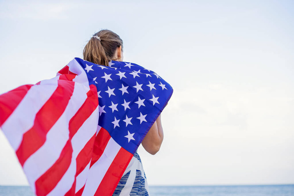 Back view of young woman holding national American flag on a ocean beach. USA Memorial day and Independence day concept. - Photo, Image
