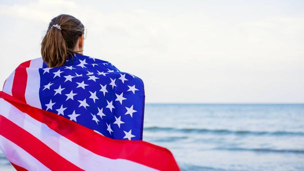 Vista trasera de una mujer joven sosteniendo la bandera nacional americana en una playa oceánica. USA Memorial day y concepto de Día de la Independencia. - Foto, imagen