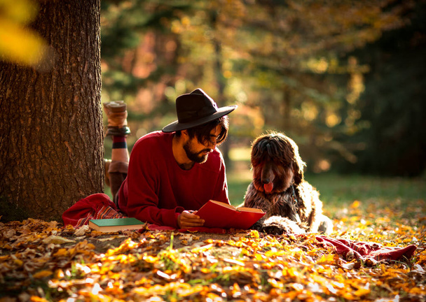 un hombre joven, moreno, con un suéter rojo y un sombrero, yace debajo de un árbol con un perro de compañía, por la tarde en un parque de otoño - Foto, Imagen