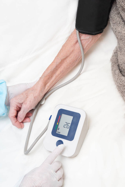 Le médecin portant des gants mesure la pression artérielle d'une femme âgée à l'aide d'un tonomètre électronique. Photo recadrée des mains. Concept d'appel d'une ambulance, examen médical à domicile - Photo, image