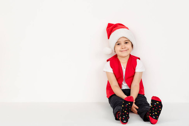 Retrato de niño feliz sentado sonriente en sombrero de Santa Claus rojo aislado sobre fondo blanco. Hermoso niño europeo de cinco años. Banner. Copiar espacio. Feliz Navidad diseño de maqueta. Tarjeta de vacaciones de Año Nuevo. - Foto, Imagen