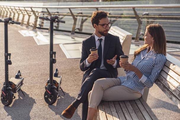 Молодий бізнесмен і бізнес-леді спілкуються, сидячи на лавці і використовуючи свої телефони під час їзди на електричних скутерів на фоні ранкового міста
. - Фото, зображення