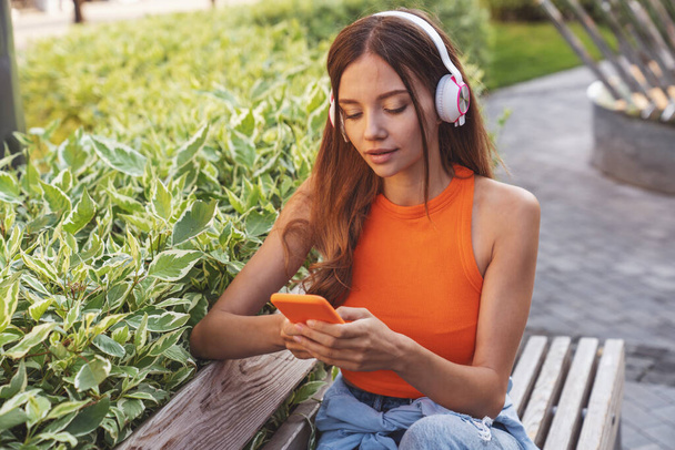 Fiatal gyönyörű lány zenét hallgat a fejhallgatón ülve a parkban padon. - Fotó, kép