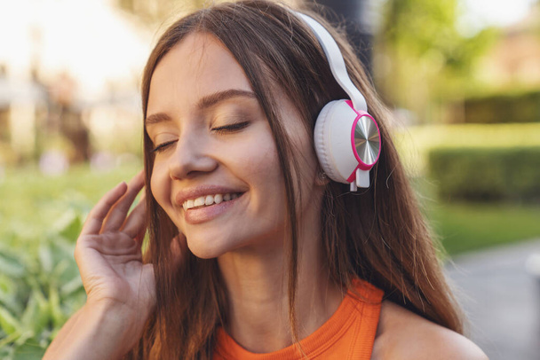 Young beautiful girl listens to music on headphones sitting on a park bench. - Foto, Imagen