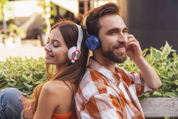Νεαρό χαμογελαστό ζευγάρι ακούγοντας μουσική στα ακουστικά κάθεται σε ένα παγκάκι πάρκο πλάτη με πλάτη. - Φωτογραφία, εικόνα
