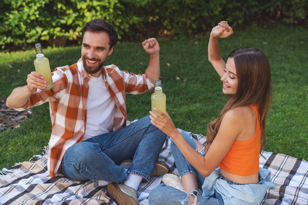 ピクニック中に公園の毛布の上に座って、ボトルからアルコールを飲む若い陽気なカップル. - 写真・画像