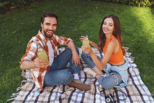 Young cheerful couple sitting on a blanket in the park during a picnic and drinking alcohol from a bottle. - Foto, imagen