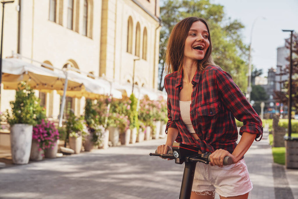 Alegre joven hermosa chica mientras monta un scooter eléctrico en una calle de la ciudad. - Foto, Imagen