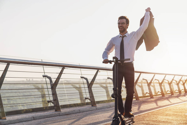 Young smiling bearded businessman in a business suit while riding an electric scooter on the background of the morning city - Photo, Image
