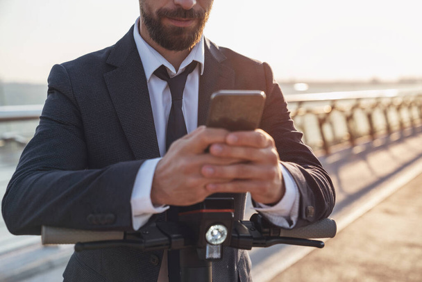 電気スクーターに乗っている間、ビジネススーツの若い陽気な髭のビジネスマンは、朝の街の背景にスマートフォンを使用しています。 - 写真・画像