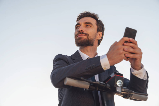 Young cheerful bearded businessman in a business suit while riding an electric scooter uses a smartphone on the background of the morning city - Photo, Image