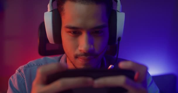 幸せなアジアの男のゲーマーは、夜の現代の家のリビングルームでスマートフォンのカラフルなネオンライトとオンラインヘッドフォン競争プレイビデオゲームを着用してください。スポーツストリーミングゲーム,遊ぶ無料オンライン. - 映像、動画