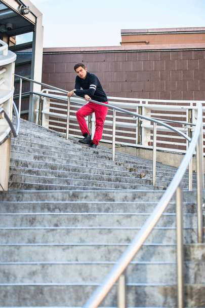 Habillé d'un pull à col châle noir et d'un jean rouge, un jeune beau garçon se tient près des balustrades d'un escalier, vous attend - Photo, image
