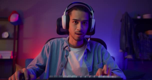 Щасливий азіатський геймер носить участь у навушниках грати у відеогру барвисті неонові вогні комп'ютер у вітальні в нічному сучасному будинку. Потокова онлайн гра Esport, концепція домашнього карантину
. - Кадри, відео