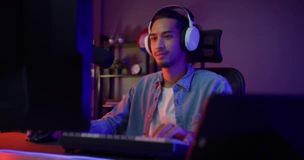 幸せなアジアの男のゲーマーは、ヘッドフォン参加プレイビデオゲームカラフルなネオンライトコンピュータを着用し、夜の現代の家のリビングルームでカメラを見て笑顔。スポーツストリーミングゲームオンライン. - 映像、動画