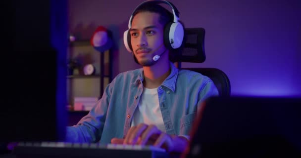 幸せなアジアの男のゲーマーは、ヘッドフォン参加プレイビデオゲームカラフルなネオンライトコンピュータを着用し、夜のモダンな家のリビングルームでタブレット上のポッドキャストを記録します。スポーツストリーミングゲームオンライン. - 映像、動画