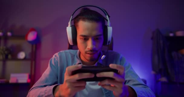 Щасливий азіатський геймер носить змагання з навушників грати в відеоігри онлайн зі смартфоном барвисте неонове світло у вітальні в нічному сучасному будинку. Esport потокова гра онлайн, Домашня карантинна діяльність
. - Кадри, відео