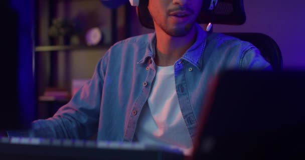 Boldog ázsiai srác játékos visel fejhallgató részvétel játék videojáték színes neon fények számítógép és rögzít egy podcast tabletta a nappaliban éjjel modern ház. Esport streaming játék online. - Felvétel, videó