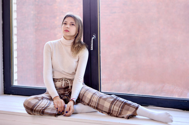 Young woman in checkered pants sits on windowsill. - Φωτογραφία, εικόνα