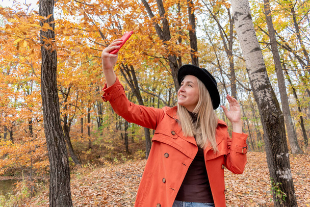 Young girl with blond hair makes a selfie on the phone while standing in the autumn forest. Concept of using technology. Copy space - Φωτογραφία, εικόνα