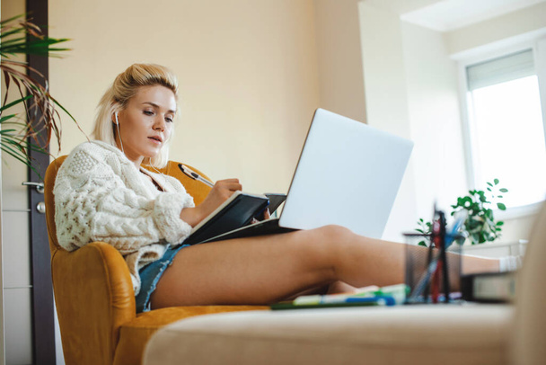 Visão inferior de uma mulher caucasiana ouvindo aulas online. De pé confortavelmente no sofá com as pernas estendidas na mesa, observando algo nela - Foto, Imagem