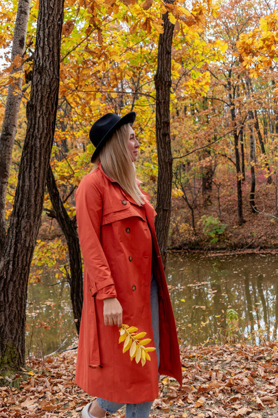 Young girl with blond hair walks in the autumn forest. Wellness concept. Orange trench coat and black hat. Copy space - Zdjęcie, obraz