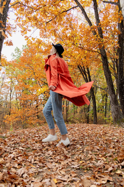 Young girl with blond hair walks in the autumn forest. Wellness concept. Orange trench coat and black hat. Copy space - Foto, imagen