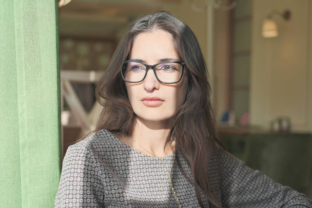 asian mixed race woman sitting in a cafe and looking aside. young beautiful businesswoman wearing glasses thinking alone. calm and thoughtful lady with long brown hair. - Foto, Imagem
