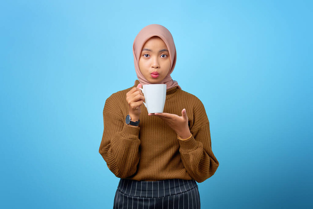 Beautiful young Asian woman holding mug and drinking tea on blue background - Foto, Imagen