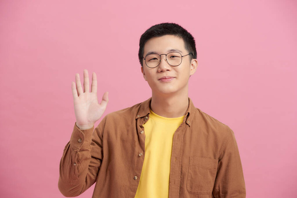 Portrait of Asian man in blue t-shirt raising hand up and welcomes with a palm and smiles - Photo, Image