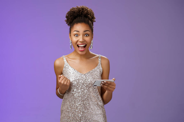 Excited charming young african-american woman lucky winning game playing smartphone standing pleased yelling happily clench fist triumphing joyfully celebrating great news received message - Фото, изображение