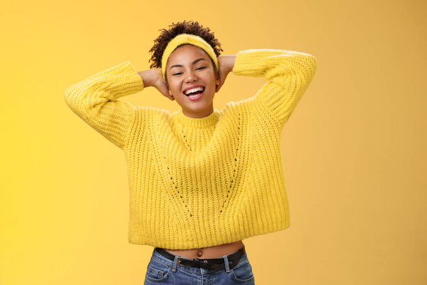 Insouciante joyeuse chanceuse étudiante afro-américaine dans le bandeau pull mettre les mains en arrière derrière la tête relaxé pose de refroidissement ayant week-end de jour-off souriant largement rire, fond jaune - Photo, image