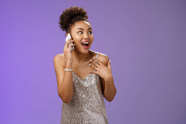 Beeindruckt gesellige afrikanisch-amerikanische gesprächige Frau im silbernen Kleid spricht Smartphone zeigt sich amüsiert beschreiben Emotionen Eindrücke nach dem Besuch genial Party, blauer Hintergrund - Foto, Bild
