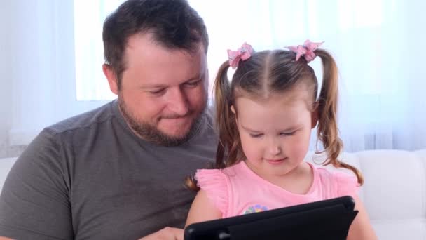 funny little girl 3 years old and her father study together on a tablet and watch a lesson online at home. Dad helps his daughter with her studies. Online school distance learning concept. - Footage, Video