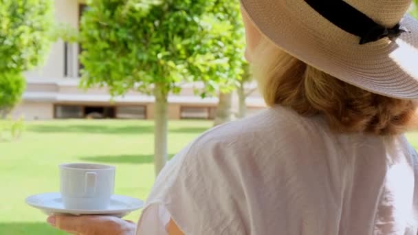 età femminile 50-55 in un cappello di paglia tiene una tazza di bevanda calda e beve caffè, seduto al mattino su una terrazza estiva in una giornata di sole. Il concetto di buongiorno, viaggio e attesa di nuovi - Filmati, video