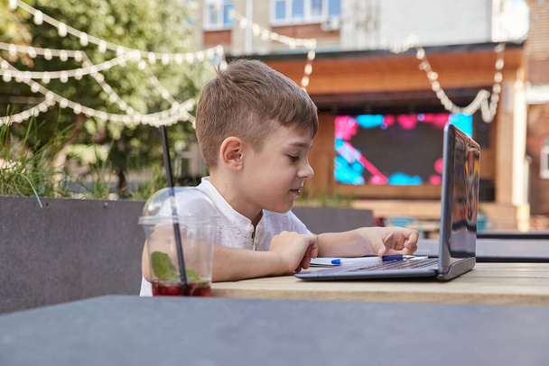 sorrindo menino caucasiano sentado à mesa na varanda do café, estudando com laptop - Foto, Imagem