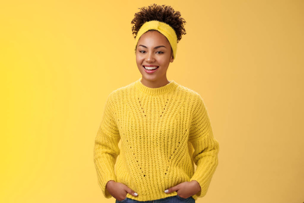 フレンドリーな発信リラックスした若いアフリカ系アメリカ人の美しい女の子の頭バンド額でセーターホールド手ポケット笑顔広く楽しい話面白いコミュニケーションを持っています,立って黄色の壁 - 写真・画像