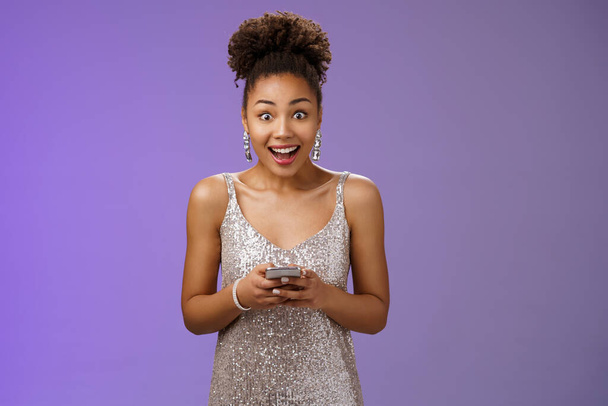 Amusé afro-amerikai lány ezüst fényes ruha kap félelmetes meglepő hír SMS-ben kiszélesíti a szemét lenyűgözte boldogan mosolygós hitetlen szerencse áll csodálkozva hold smartphone - Fotó, kép