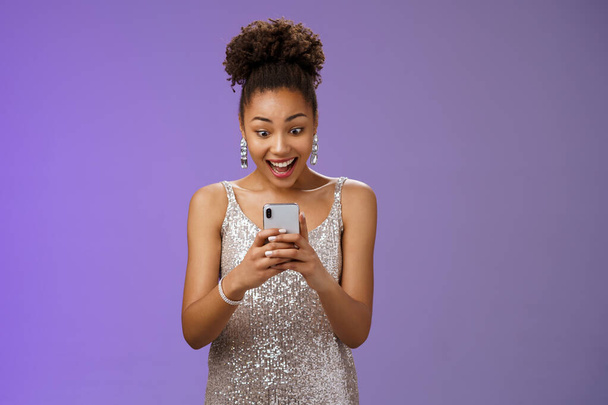 Good-looking amazed african-american woman in silver shiny dress smiling amused reading awesome news message smartphone look phone display astonished happy blue background - Photo, Image