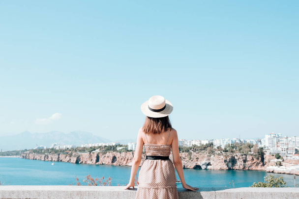 A young female tourist in a dress and hat on the viewpoint against the background of the city Antalya. Female tourist enjoying the cityscape . - Photo, Image