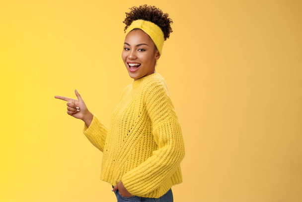 Нахальный милый женский африканско-американская девушка 20 лет в свитере ободок улыбаясь нахальный флирт смотреть камера поворачивая стоя профиль указывая левый интродуцированный удивительный новый продукт показывая путь, желтый фон - Фото, изображение
