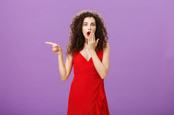 Portrait of surprised and amazed charming elegant lady with evening make-up in red dress covering opened mouth from shock and amazement pointing left curious and questioned over purple background - Photo, Image