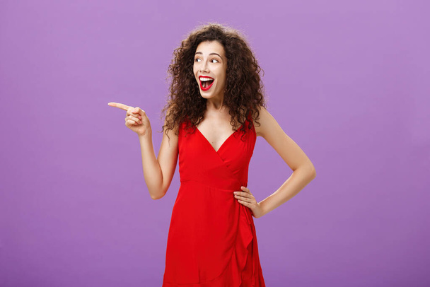 Amused attractive european female with evening makeup in luxurious red dress and curly hairstyle laughing out loud entertained and happy pointing and gazing left intrigued over purple background - Zdjęcie, obraz