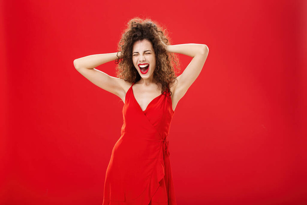 Girl just wanna have fun. Crazy adult caucasian woman with curly hairstyle in elegant dress holding hands on head yelling out loud with closed eyes and intense expression over red background - Photo, Image