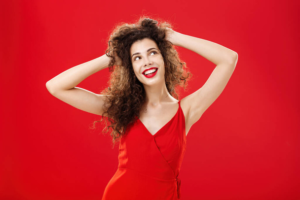Dreamy carefree young european female with curly hairstyle and evening make-up touching hairstyle pleased with result after visiting salon looking up dreaming or imaging perfect date in red dress - Fotoğraf, Görsel