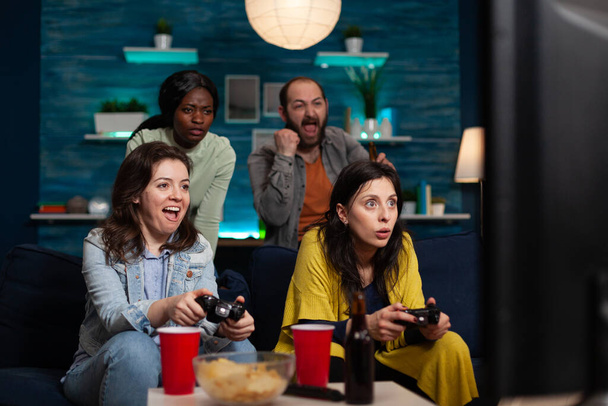 Emocionadas amigas sentadas en sofás ganando videojuegos usando joystick de juegos - Foto, imagen