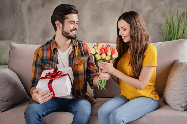 Photo of attractive beautiful young para nosić casual stroje uśmiechnięty prezentując tulipany pudełko prezentów wewnątrz domu pokój - Zdjęcie, obraz