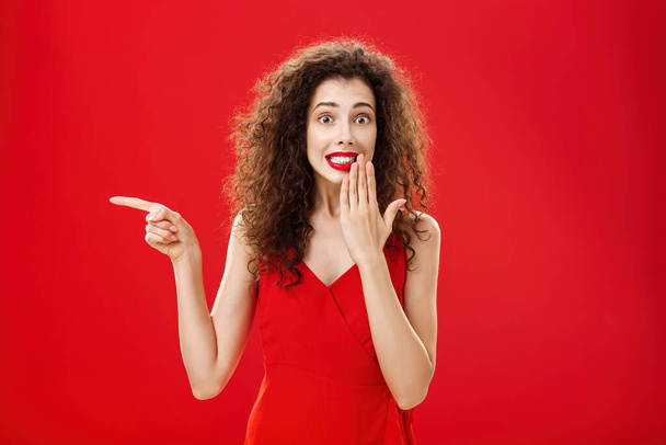 Troubled cute and tender silly woman with curly hairstyle in red stylish evening dress saying oops smiling with guilty look covering mouth with palm while pointing left over studio background - Фото, изображение