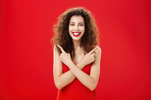 Γυναίκα ικανοποιημένη με κάθε απόφαση αισθάνεται ανέμελη και ευτυχισμένη στέκεται στο κομψό κόκκινο φόρεμα βράδυ με σγουρά μαλλιά σταυρώνουν τα χέρια στο στήθος δείχνοντας αριστερά και δεξιά ενθουσιασμένοι με τις επιλογές - Φωτογραφία, εικόνα