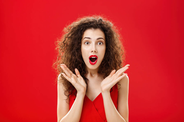 Waist-up shot of amused and astonished good-looking young elegant female with curly hairstyle in red lipstick raising palms in surprise opening mouth and looking at camera intrigued and shocked - Photo, Image